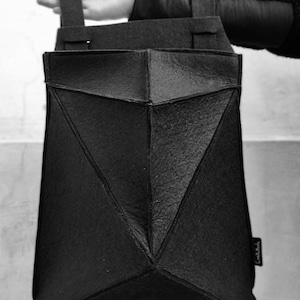 May include: Black geometric tote bag with a textured surface. The bag has a unique origami-like design with angular panels and two shoulder straps. A small tag with text is visible on the side.