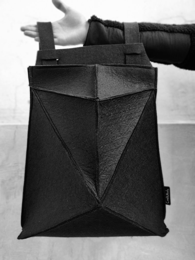 May include: Black geometric tote bag with a textured surface. The bag has a unique origami-like design with angular panels and two shoulder straps. A small tag with text is visible on the side.
