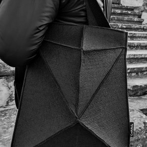 May include: Black geometric tote bag with a textured surface, carried over the shoulder. The bag has a unique angular design and a long strap. The bag is being carried by a person wearing a black jacket.