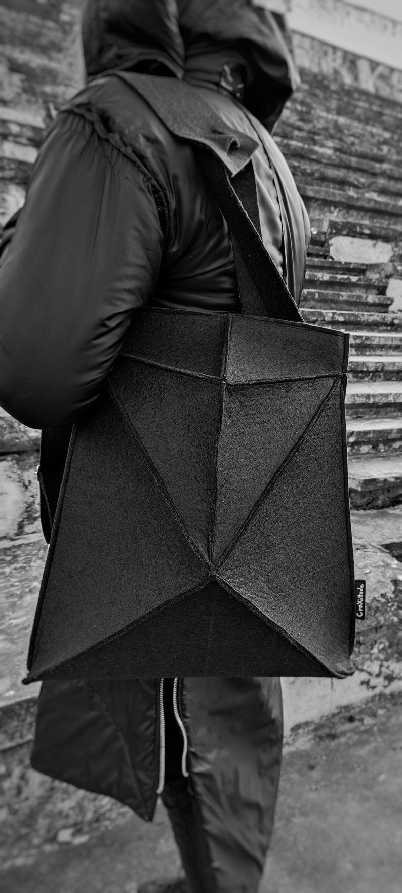 May include: Black geometric tote bag with a textured surface, carried over the shoulder. The bag has a unique angular design and a long strap. The bag is being carried by a person wearing a black jacket.