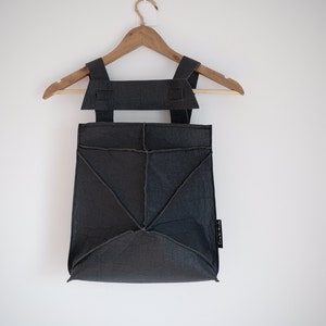 May include: A dark gray backpack with geometric stitching and adjustable straps hangs on a wooden hanger. The bag has a unique, angular design and a small tag on the side. The background is a plain white wall.