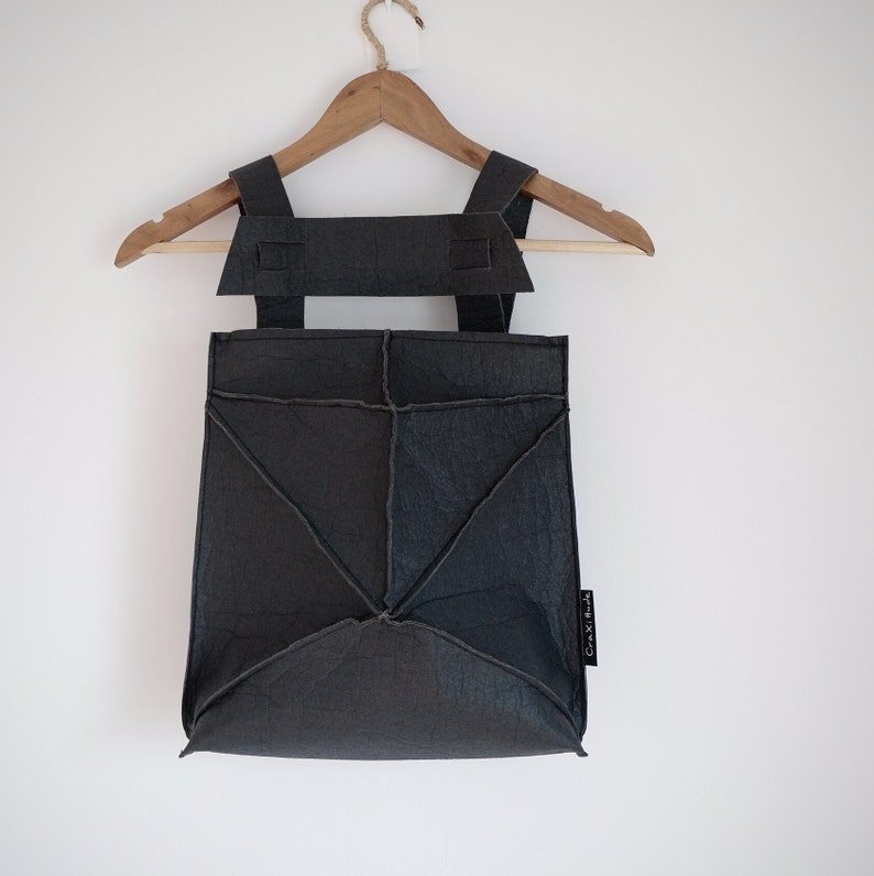 May include: A dark gray backpack with geometric stitching and adjustable straps hangs on a wooden hanger. The bag has a unique, angular design and a small tag on the side. The background is a plain white wall.