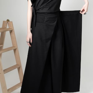 Könnte beinhalten: Ein schwarzer Wickelrock und ein passendes Oberteil. Das Oberteil hat ein asymmetrisches Design mit drapierter Schulter und Bindedetail. Der Rock ist lang und weit geschnitten. Das Outfit wird mit schwarzen Stiefeln kombiniert. Eine Holzleiter ist im Hintergrund.