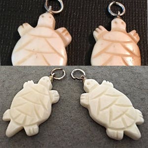 Könnte beinhalten: Vier weiße, geschnitzte Schildkröten-Anhänger mit goldfarbenen Sprungringen. Die Schildkröten haben detaillierte Schnitzereien auf ihren Panzern und sind ein beliebtes Symbol für Glück und Langlebigkeit.
