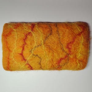 Puede incluir: Una pastilla de jabón rectangular de fieltro hecha a mano. El jabón es principalmente naranja con patrones rojos y amarillos en forma de vetas. La textura parece suave y ligeramente borrosa, sugiriendo un producto natural y artesanal. Mide unos 10 cm.