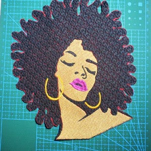 May include: Embroidered patch featuring a Black woman with closed eyes, gold skin, and vibrant pink lips. The patch has black textured hair with a red outline, and gold hoop earrings. The design is on a green cutting mat.