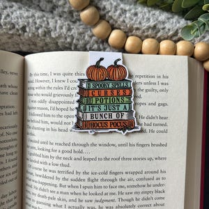 May include: A Halloween-themed bookmark with a stack of books featuring the text "Spooky Spells", "Curses", "Potions", "It's Just a Bunch of", and "Hocus Pocus". The bookmark is decorated with two orange pumpkins.