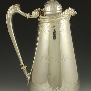 May include: A silver coffee pot with an ornate handle and a lid with a finial. The pot is engraved with the name "JON. SAY."