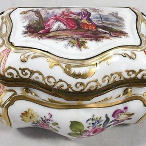 May include: A white porcelain box with a gold lid and floral designs. The lid features a painted scene of two figures in a landscape.