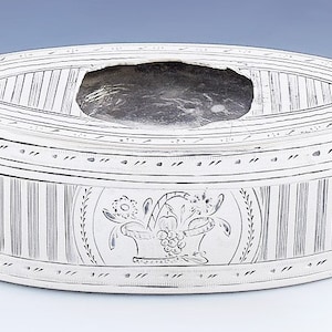 Può includere: Una scatola ovale in argento con un design inciso di un cesto di fiori. La scatola ha un coperchio con un'apertura circolare al centro.