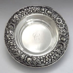 May include: A silver serving dish with a floral design around the rim. The dish has a monogram of the letter "C" in the center.