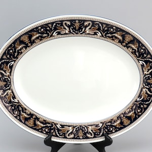 May include: An oval serving platter with a white centre and a black and gold border. The border features a repeating pattern of stylised floral and leaf designs.