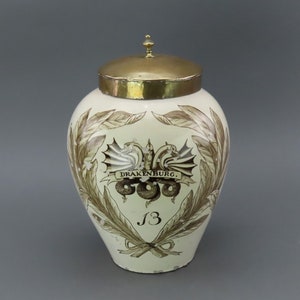 Pode incluir: Um pote de cerâmica branca com uma tampa de latão. O pote é decorado com um desenho marrom e branco de um dragão e uma coroa de folhas. O texto "DRAKENBURG." está impresso no pote.