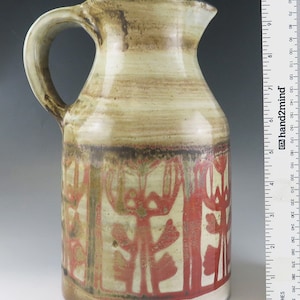 Puede incluir: Una jarra de cerámica con un esmalte marrón, blanco y rojo. La jarra tiene un diseño floral estilizado y un asa.