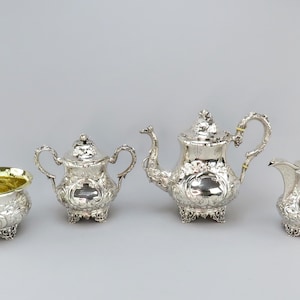 Puede incluir: Un juego de té de plata con tetera, jarra de crema, azucarero y un tazón pequeño. El juego está decorado con intrincados diseños florales.