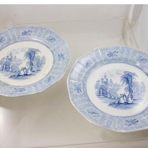 May include: Two blue and white porcelain plates with a decorative border and a central image of a castle and trees.