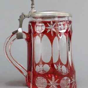 May include: A red glass beer stein with a cut glass design and a pewter lid. The stein has a handle and is decorated with a starburst pattern.