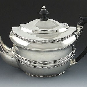 Może przedstawiać: Srebrny czajnik z czarnym drewnianym uchwytem i gałką. Czajnik ma zaokrąglony korpus z trzema ozdobnymi paskami wokół środka.