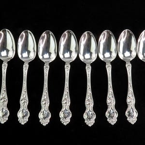 May include: A set of twelve silver teaspoons with ornate handles. The spoons are arranged in two rows of six, with the handles facing outwards.