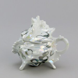 Può includere: Una teiera in porcellana bianca con un coperchio a forma di conchiglia. La teiera ha un manico e tre gambe. La teiera è decorata con una glassa iridescente bianca e nera.
