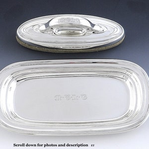May include: A silver butter dish with a lid. The dish has a rounded rectangular shape with a raised rim. The lid has a handle and is engraved with the text "M-G-L".