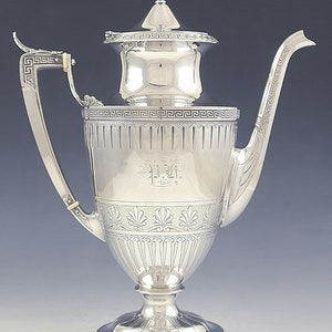 May include: A silver teapot with a decorative design and a Greek key pattern around the base. The teapot has a lid with a finial and a spout with a curved handle.