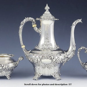 May include: A silver tea set with a teapot, creamer, and sugar bowl. The set is ornately decorated with floral and scroll designs. The teapot has a domed lid and a spout with a curved handle. The creamer and sugar bowl have matching handles and feet.