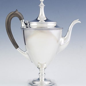 May include: A silver teapot with a wooden handle and a square base. The teapot has a tall, slender spout and a lid with a finial.