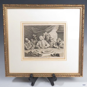 May include: A black and white engraving of a group of men seated around a table, with a plate of food in the center. The engraving is framed in a gold frame.