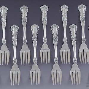 Könnte beinhalten: Eine Sammlung silberner Gabeln, angeordnet auf grauem Untergrund. Die Gabeln haben verzierte Griffe mit floralem Design und drei Zinken. Das Bild zeigt verschiedene Gabelgrößen, was auf ein komplettes Set für ein formelles Abendessen hindeutet.