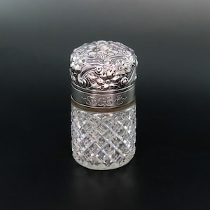 May include: A silver and glass bottle with a decorative floral design on the lid. The bottle is engraved with the letters "J & C".