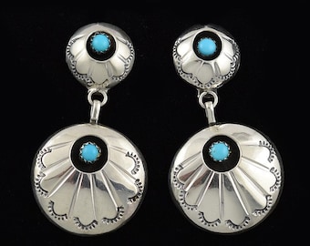 May include: A pair of silver dangle earrings featuring turquoise stones. Each earring has two round, silver discs with a turquoise stone set in the centre of each disc. The discs are decorated with a scalloped edge.