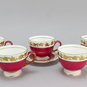 Puede incluir: Juego de cinco tazas y platillos vintage. Las tazas y los platillos tienen un interior blanco, un exterior rojo texturizado y un diseño de hojas doradas. Las tazas tienen asas doradas y se asientan sobre una base blanca.