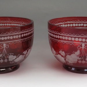 May include: Two red glass bowls with white etched designs. The bowls have a scalloped rim and a floral pattern with a ribbon.