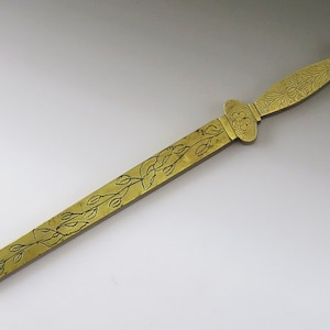 Könnte beinhalten: Ein goldfarbener Brieföffner aus Metall mit floralem Design. Der Griff ist schwertförmig und hat ein dekoratives Blumenmuster.