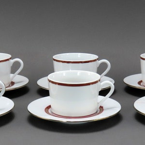 May include: Six white teacups and saucers. Each cup and saucer has a dark red band around the rim and a gold trim. The cups have a curved handle. The saucers are round.