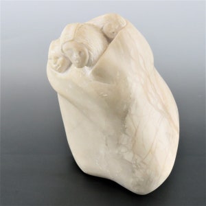May include: A white alabaster sculpture of three figures huddled together under a blanket. The sculpture is carved in a realistic style and depicts the figures in a protective embrace.