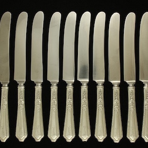 May include: A set of 12 silver dinner knives with ornate handles. The knives have a polished finish and are arranged in a row.