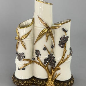 May include: A white ceramic vase shaped like three bamboo stalks with gold and silver painted details. The vase has three openings for flowers and a gold and brown base.