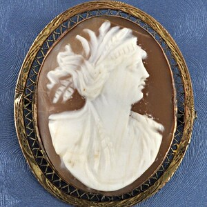 Puede incluir: Un broche de camafeo con un perfil tallado de una mujer con un tocado de plumas, engastado en un marco de metal dorado con un delicado diseño de alambre.