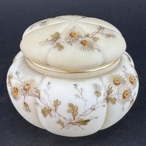 May include: A white ceramic jar with a lid, decorated with a floral pattern in shades of brown and yellow. The jar has a rounded shape and a gold trim around the lid.