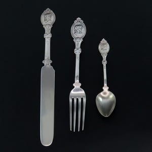 May include: A set of three silver-plated utensils, including a butter knife, fork, and spoon. Each utensil features an ornate handle with a decorative design.