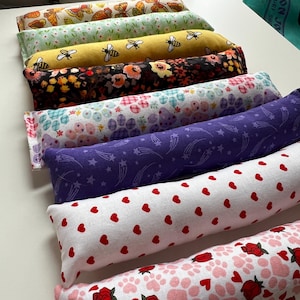 Può includere: Sette giocattoli per gatti in tessuto colorato a forma di cilindro lungo. I giocattoli sono impilati, con il giocattolo superiore di colore rosa chiaro con un'impronta di zampa bianca e un motivo di rose. Il giocattolo successivo è bianco con cuori rossi, seguito da un giocattolo viola con stelle bianche, un giocattolo bianco con fiori rosa e bianchi, un giocattolo nero con fiori colorati, un giocattolo giallo con api e un giocattolo verde con farfalle.