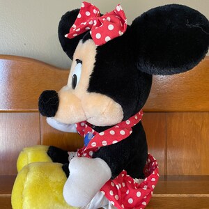Pu&ograve; includere: Un giocattolo di peluche di Minnie Mouse con un vestito rosso a pois bianchi e un fiocco rosso a pois bianchi. Ha le orecchie nere e un viso giallo. &Egrave; seduta con le gambe incrociate.