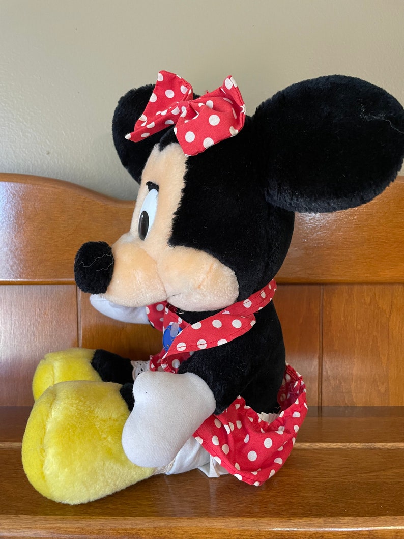 Pu&ograve; includere: Un giocattolo di peluche di Minnie Mouse con un vestito rosso a pois bianchi e un fiocco rosso a pois bianchi. Ha le orecchie nere e un viso giallo. &Egrave; seduta con le gambe incrociate.