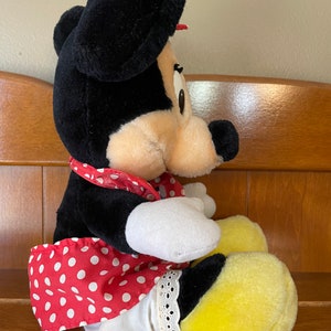 Pu&ograve; includere: Un giocattolo di peluche di Minnie Mouse che indossa un vestito rosso con pois bianchi e scarpe gialle. Ha un fiocco nero sulla testa e un nastro rosso intorno al collo.