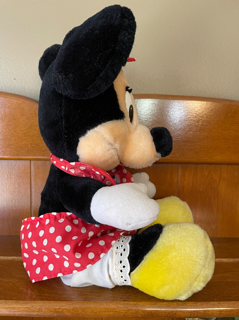 Pu&ograve; includere: Un giocattolo di peluche di Minnie Mouse che indossa un vestito rosso con pois bianchi e scarpe gialle. Ha un fiocco nero sulla testa e un nastro rosso intorno al collo.