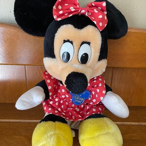 Pu&ograve; includere: Un giocattolo di peluche di Minnie Mouse che indossa un vestito rosso con pois bianchi e un fiocco rosso con pois bianchi. Il giocattolo ha un'etichetta blu Disney Original sul petto.