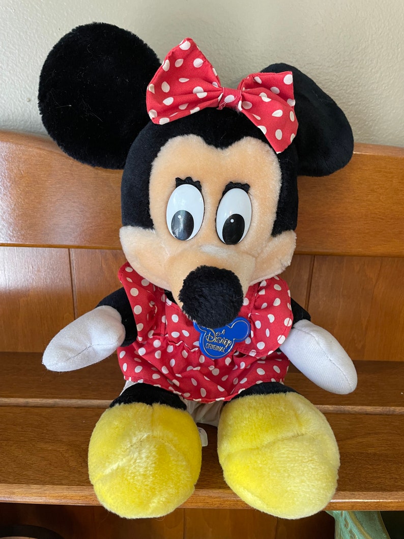 Pu&ograve; includere: Un giocattolo di peluche di Minnie Mouse che indossa un vestito rosso con pois bianchi e un fiocco rosso con pois bianchi. Il giocattolo ha un'etichetta blu Disney Original sul petto.
