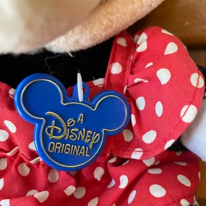 Pu&ograve; includere: Un'etichetta di plastica blu a forma di testa di Topolino con il testo "A Disney Original" in lettere dorate. L'etichetta &egrave; attaccata a un vestito rosso con pois bianchi.